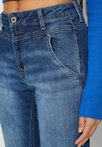Close-up of midsection wearing blue faded jeans and a bright blue long-sleeve top, with hand resting by the thigh.
