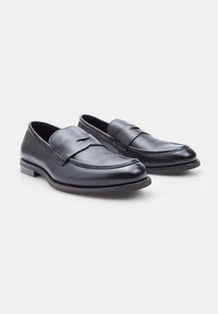 Mocasines de cuero negro con un acabado texturizado, que presentan un acento cosido en la parte delantera y un diseño de punta redondeada y suave. Suela plana y resistente.