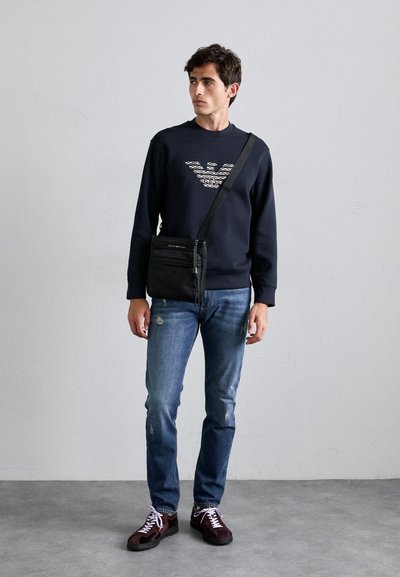 Jonge man in een marineblauw sweatshirt met logo, blauwe versleten jeans, donkere sneakers en een zwarte schoudertas, staande op een grijze vloer en muur.
