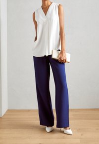 Haut blanc sans manches, pantalon large bleu marine, et talons pointus blancs. Tient une petite pochette dorée. Fond en parquet en bois.