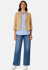 Veste beige cropped avec un col cranté, portée sur une chemise à rayures bleues et blanches à boutons, associée à un jean en denim bleu à jambes larges et des baskets.