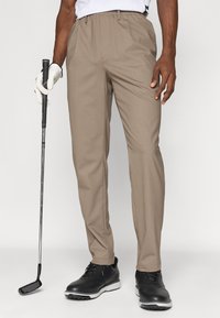 Pantaloni de golf bej, cu o textură netedă și un design cu fața plată. Împerechează cu pantofi de golf negri, ținând un club de golf cu o mână purtând o mănușă.