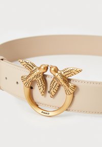 Beige leren riem met een decoratieve gouden gesp in de vorm van twee vogels. Gedetailleerde graveringen op de vogels voegen textuur toe.