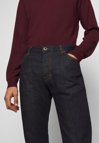 Man som bär mörkblå jeans med orange sömmar och en vinröd långärmad tröja, med handen i fickan mot en neutral bakgrund.