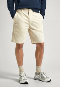 Beige bomullsshorts med rak klipp, med sidofickor och knäppning fram, kombinerat med grå och vita sneakers samt beige strumpor.