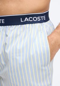 Homme portant un boxer Lacoste à rayures bleu clair et jaunes avec une ceinture marine, main dans la poche, taille et partie du torse visibles.
