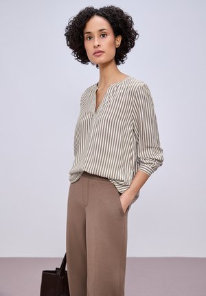 Kvinde med krøllet hår iført beige og hvidstribet bluse, stoppet ned i højtaljede taupebukser, står med den ene hånd i lommen.