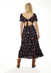 Maxi dress with a black floral print, puff sleeves, open back with a tied closure, ruffled hem, and paired with brown ankle boots.