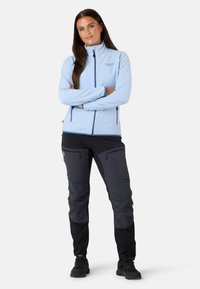 Jasnoniebieska polarowa kurtka z wysokim kołnierzem i zamykanymi na zamek kieszeniami, w połączeniu z czarnymi spodniami outdoorowymi z kontrastowymi sekcjami tkaniny i zamkami.