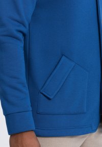 Sudadera azul con un bolsillo tipo canguro, puños acanalados y una textura de tela suave. El diseño es sencillo y ajustado.