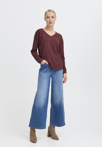 Burgunderfarbene Langarm-V-Ausschnitt-Bluse kombiniert mit weit geschnittenen blauen Jeans. Das Outfit zeichnet sich durch eine lockere Passform und einen subtilen Verblassungsbereich am Saum der Jeans aus.