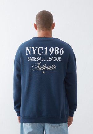 Mężczyzna stojący tyłem, ubrany w granatową bluzę z białym napisem "NYC1986 Baseball League Authentic" oraz jasnoniebieskie dżinsy.