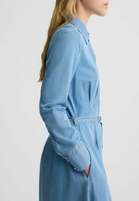 Hellblaues Denimkleid mit langen Ärmeln, Knopfmanschetten, Kragen und tailliertem Schnitt. Glatte Textur, gerades Design, knielang.
