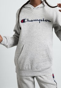 Šedá mikina s kapucí a přední kapsou. Obsahuje logo "Champion" v modré a červené barvě. Měkká texturovaná tkanina s žebrovanými manžetami a spodním lemem.