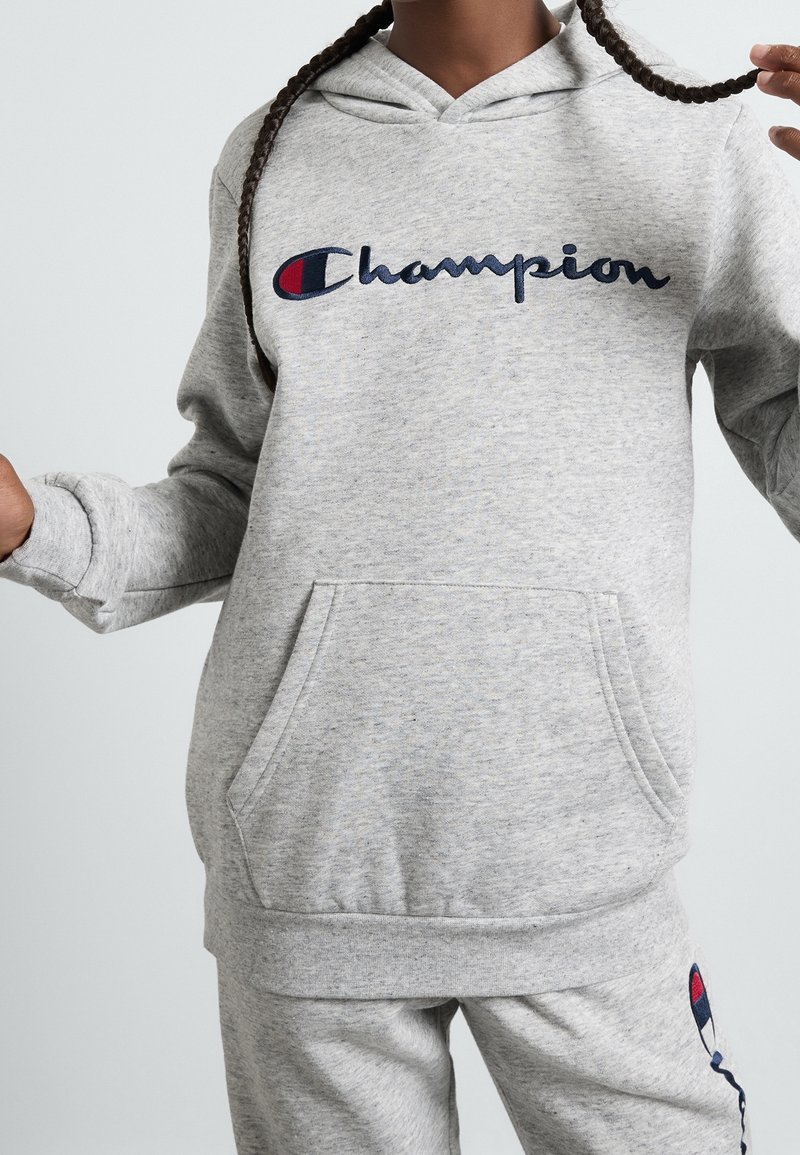 Šedá mikina s kapucí a přední kapsou. Obsahuje logo "Champion" v modré a červené barvě. Měkká texturovaná tkanina s žebrovanými manžetami a spodním lemem.