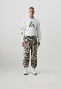 Hvit langermet skjorte med grønne grafiske design, kombinert med kamuflasje cargo bukser. Hvite sneakers fullfører antrekket.