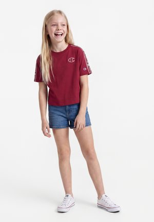 Jeune fille aux longs cheveux blonds porte un T-shirt rouge Champion, un short en jean bleu et des baskets blanches, souriante et debout sur un fond blanc.