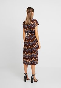 Robe à motif chevron marron avec une taille à nouer et des manches courtes, faite en tissu léger, portée avec des sandales à talons noirs.