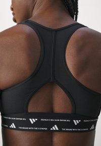 Zwarte sportbeha met racerback ontwerp, textuur stof en een brede elastische band met een wit merklogo en driehoekige accenten.