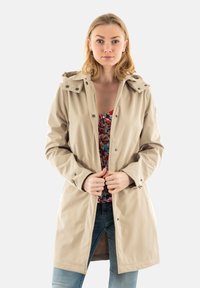 Chaqueta impermeable beige con capucha, botones a presión y bolsillos. Lleva puesta una blusa floral colorida y se combina con vaqueros azules. Textura suave.