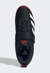 Sort atletisk sko med en tekstureret overdel, hvide striber og en velcrolukning. Har røde detaljer på sålen og hælen.