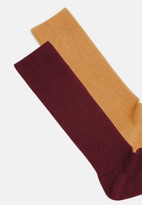 Twee geribbelde sokken: de ene ambergeel, de andere diep bordeauxrood. Elke sok heeft een gladde textuur met contrasterende kleuren en een effen ontwerp.