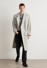Abrigo de lana gris claro con solapa con muesca, llevado sobre una camiseta gris, combinado con jeans oscuros y zapatillas negras. Textura plana, silueta larga.