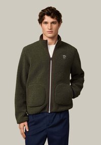 Vihreä fleecetakki vetoketjulla, kahdella suurella etutaskulla, kontrastivärisillä ruskeilla reunoilla ja logolla vasemmassa rinnassa. Pehmeä pinta.