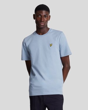 Jeune homme portant un t-shirt à manches courtes bleu clair avec un petit logo d'oiseau jaune sur la poitrine, se tenant devant un fond gris uni.