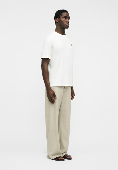 Άνδρας που φοράει μπεζ παντελόνι με φαρδιά μπατζάκια, λευκό κοντομάνικο πουκάμισο με μικρό λογότυπο στο στήθος και μαύρα σανδάλια, μπροστά σε απλό φωτεινό φόντο.