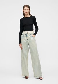 Γυναίκα στέκεται με το χέρι στην τσέπη, φορώντας μαύρο μακρυμάνικο τοπ, ανοιχτόχρωμα τζιν με φαρδιά παντελόνια και μαύρες ψηλοτάκουνες ανοικτές γόβες σε απλό φόντο.