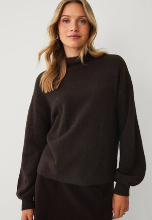Blonde woman with loose hair wears dark brown long-sleeve sweater and matching skirt, standing against plain light background.