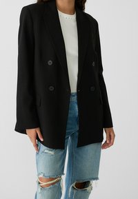 Black double-breasted blazer with notch lapels, textured fabric, and two front pockets worn over a white top and distressed light blue jeans.