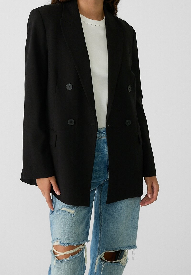 Black double-breasted blazer with notch lapels, textured fabric, and two front pockets worn over a white top and distressed light blue jeans.
