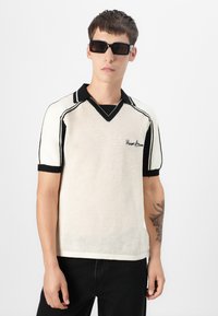 Gestricktes Poloshirt in Creme mit schwarzen Akzenten. Verfügt über einen V-Ausschnitt, kurze Ärmel und ein strukturiertes Muster. Mit einem gestickten Logodetail versehen.