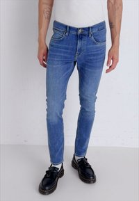 Jeans de mezcla en azul claro, de corte slim-fit, con un diseño clásico de cinco bolsillos, fabricados en denim, con un ligero desgastado, combinados con zapatos negros y calcetines blancos.
