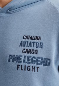 Plava dukserica s vezom koja prikazuje tekst "CATALINA AVIATOR CARGO PME LEGEND FLIGHT" u kontrastnoj tamnoplavoj i crnoj boji. Mekani tkaninski tekst.