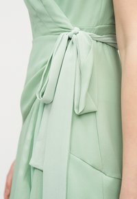 Κοντινό πλάνο ζώνης με φιόγκο σε ανοιχτό πράσινο φόρεμα που φορά ένα άτομο, τονίζοντας την υφή του υφάσματος και τη λεπτομέρεια του κόμπου.