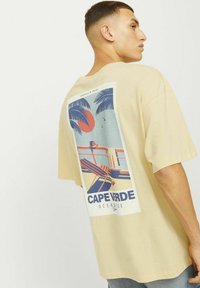 Beige oversized t-shirt met een kleurrijke graphic print van palmbomen en een zonsondergang. Korte mouwen en een ronde halslijn.