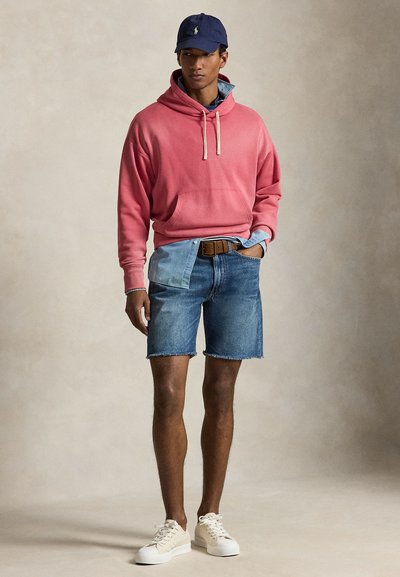 Férfi rózsaszín kapucnis pulóvert visel farmerruhával, tengerészkék sapkával, kék farmer rövidnadrággal nyers szegéllyel, barna övvel és fehér tornacipővel, egyszínű háttér előtt állva.