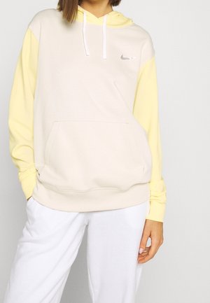 Vrouw draagt een tweekleurige beige en lichtgele Nike hoodie met witte trekkoorden, gecombineerd met een witte broek, staand tegen een effen achtergrond.
