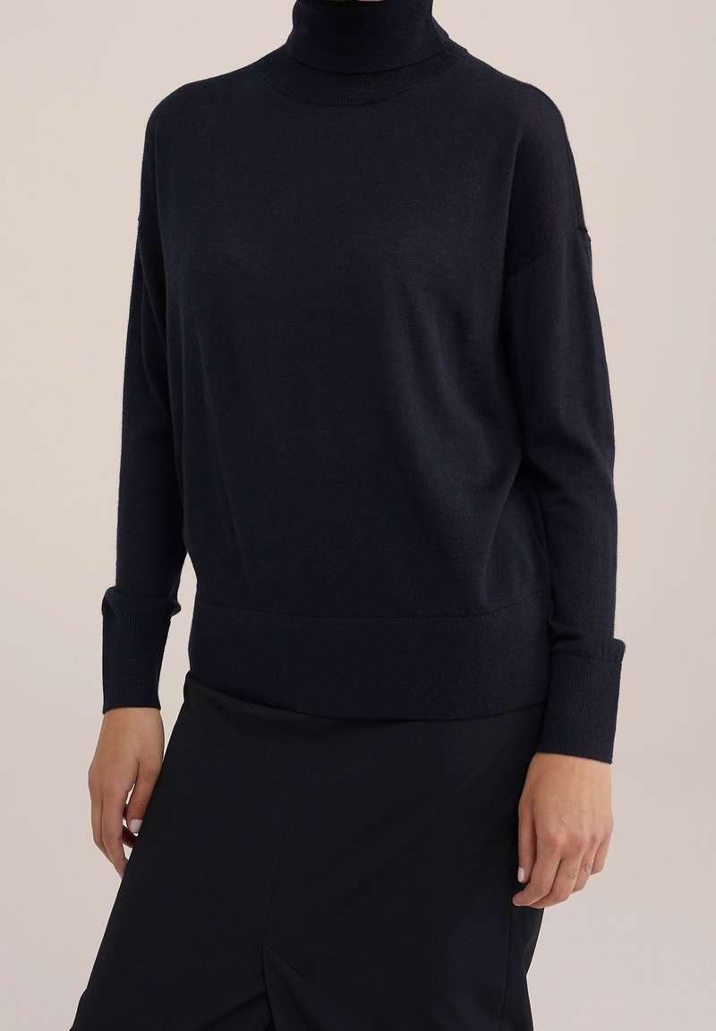 Kvinna klädd i en svart polotröja med långa ärmar och svart kjol mot en enkel beige bakgrund.