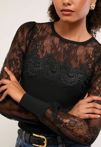 Fekete, hosszú ujjú felső, áttetsző csipke betéttel, virágmintákkal, textúrált ujjal és kerek nyakkal. Hímzett részletekkel díszítve.