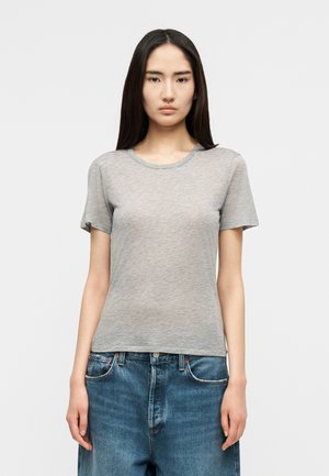 Tânără cu păr lung și închis la culoare, purtând un tricou gri simplu cu mâneci scurte și blugi albaștri din denim, stând în fața unui fundal alb.