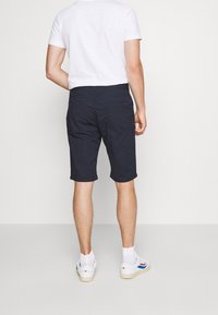 Shorts bleu marine avec un motif texturé, coupe droite, et deux poches arrière, portés avec un t-shirt blanc et des baskets blanches.