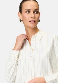 White button-up shirt with navy vertical stripes, featuring a classic collar, long sleeves, and cuff details. Smooth texture, light fabric.