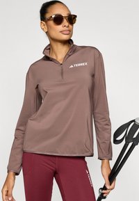 Barna, hosszú ujjú, fél cipzáras pulóver Terrex logóval; bordó leggingszel párosítva. A modell trekkingbotokat tart fekete markolatokkal.