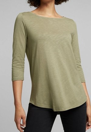 Olivgrünes Langarmshirt mit Bootsausschnitt, lockerem Schnitt und weichem, strukturiertem Stoff. Der Saum ist leicht geschwungen. Kombiniert mit schwarzer Hose.