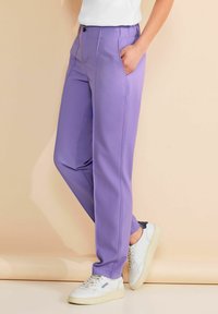 Persoon die lila rechte broek met zakken en witte sneakers draagt, staand tegen een beige achtergrond.