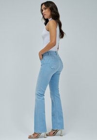 Vaqueros de campana azul claro hechos de mezclilla, con cintura alta, bolsillos laterales y un sutil efecto de lavado. Combinados con una camiseta sin mangas blanca y tacones.